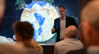 Foredragsholder peker og forklarer foran et stort kart over Arktis på en skjerm, med publikum sittende i forgrunnen.