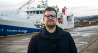 En person med kort hår og briller står utendørs på en kai. I bakgrunnen ligger et stort skip fortøyd ved havn, med kran og overbygg synlig. Personen har på seg en mørk jakke, og det er overskyet vær.
