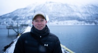 En smilende mann i vinterklær står på en brygge ved vannet, med snødekte fjell i bakgrunnen og lett snøfall rundt seg.