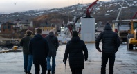 Fem personer går langs en kai i et lite kystsamfunn med snødekte fjell i bakgrunnen og industrielt utstyr rundt seg.