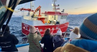 Turister i varme klær står på dekk av en båt og tar bilder av en rød fiskebåt på havet i arktisk landskap ved solnedgang.