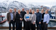En gruppe på ti personer står samlet ved en havn med båter og snødekte fjell i bakgrunnen. En kvinne i midten holder en rapport, og alle vender mot kameraet.