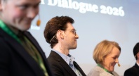 Tre personer med hodesett sitter på en scene under et arrangement, med teksten «Dialogue 2026» synlig i bakgrunnen.