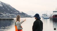 To personer står ved en havn og ser ut over vannet mot båter og snødekte fjell.
