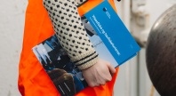 Person i oransje arbeidsklær holder en blå brosjyre med tittelen ‘Yrkesfiske og havbrukslære’, stående utendørs