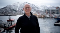 Mann med briller står ved en havn, med båter og snødekte fjell i bakgrunnen.