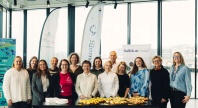 En gruppe på rundt 14 personer står samlet bak et bord med småretter i et lyst lokale, smilende mot kamera, med bannere i bakgrunnen.