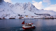 Fiskebåt på vei inn mot et snødekt kystsamfunn omgitt av bratte fjell i nordnorsk vinterlandskap.