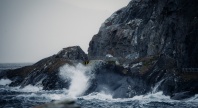 Bølger slår mot en bratt og værutsatt kystvei langs havet. En bil kjører på den smale veien mellom mørke klipper og opprørt sjø.