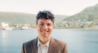 Mann iført dressjakke, smiler til kamera. Hav i bakgrunnen.