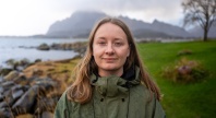 Therese Hugstmyr Woie står ved en steinete kystlinje med havet og fjell i bakgrunnen. Hun har på seg en grønn jakke og ser mot kameraet.